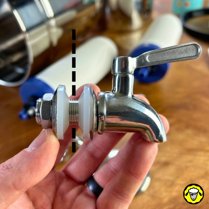 Image montrant le robinet inox d'un filtre a gravité Pure-Filters, équipé de ses deux joints en caoutchouc, ses deux rondelles en inox et son écrou de serrage inox. Une ligne en pointillé schématise l'emplacement de la paroi de la cuve inox sur laquelle ce robinet est destiné.