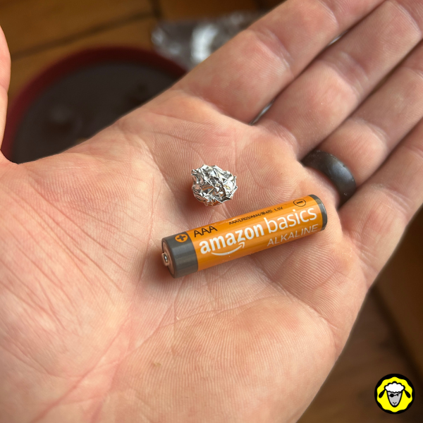 Une main tenant une pile AAA et une boule d’aluminium, montrant une astuce de remplacement pour une pile AA.