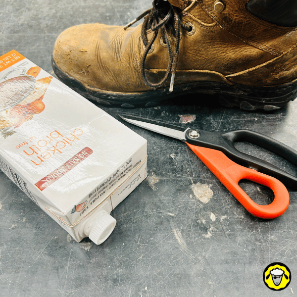 Une paire de chaussures de randonnée, des ciseaux, et une brique de carton (type bouillon) placés sur une surface, montrant les matériaux nécessaires pour fabriquer une semelle isolante.