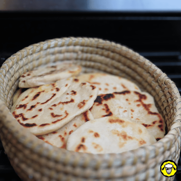 pain-maison-facile-cuit-a-la-poele-1 Un panier en osier rempli de petits pains plats dorés avec des marques de cuisson, empilés et prêts à être dégustés.