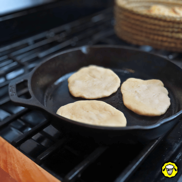 pain-maison-facile-cuit-a-la-poele-11 Trois pains plats crus sont disposés dans une poêle en fonte chauffée sur une cuisinière, prêts à être cuits.