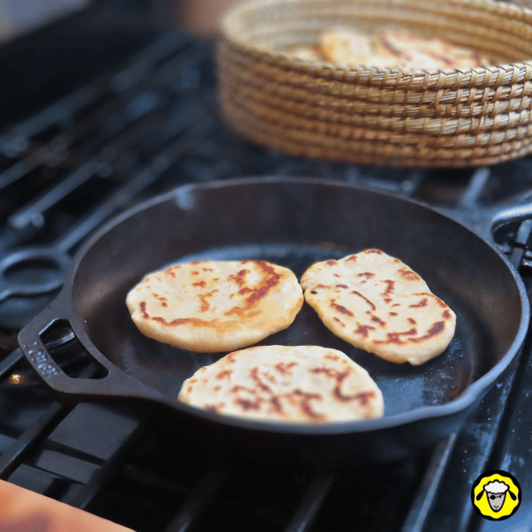 pain-maison-facile-cuit-a-la-poele-12 Trois pains plats dorés avec des marques de cuisson sont en train de cuire dans une poêle en fonte, avec un panier en osier contenant d’autres pains en arrière-plan.