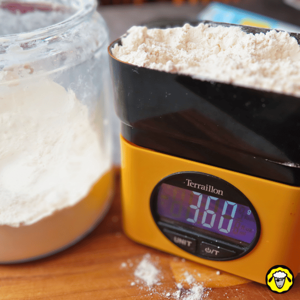 pain-maison-facile-cuit-a-la-poele-2 Une balance de cuisine numérique affichant 360 g de farine, avec un pot de farine à côté, sur un plan de travail en bois.