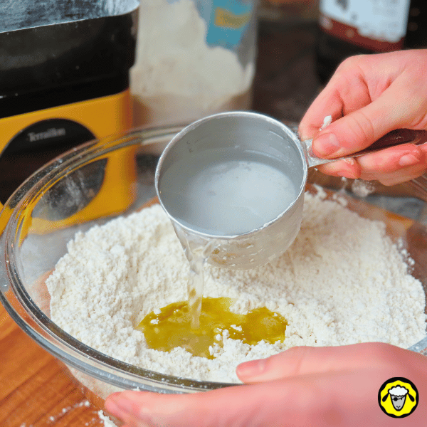 pain-maison-facile-cuit-a-la-poele-3 Une main verse de l’eau dans un mélange de farine et d’huile dans un bol en verre, préparant la pâte à pain.