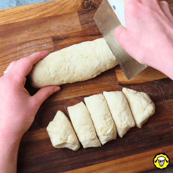 pain-maison-facile-cuit-a-la-poele-6 Une pâte roulée en boudin est coupée en portions égales sur une planche en bois à l’aide d’un coupe-pâte.
