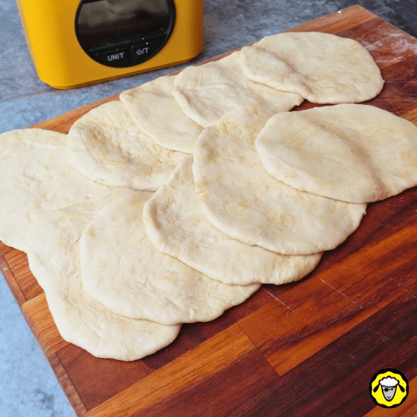 pain-maison-facile-cuit-a-la-poele-9 Plusieurs disques de pâte prêts à cuire sont disposés sur une planche en bois, avec une balance de cuisine en arrière-plan.