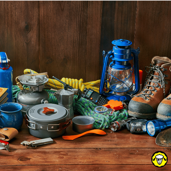 Photo d’un ensemble de matériel de camping et de survie posé sur une table en bois : réchaud, corde, lampe, bottes, popote, outils multifonction. Image représentative du kit de survie civil, de l’équipement de base pour autonomie ou d’un DOS (domicile optimisé survivaliste) bien pensé.