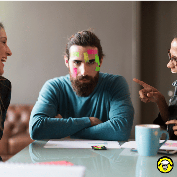 Homme barbu assis en réunion, le visage couvert de post-its, pendant que des collègues rient. Représente la marginalisation ou l’incompréhension ressentie par certains survivalistes ou autonomes dans un environnement professionnel ou social. Idéal pour illustrer l’isolement du préparé discret.