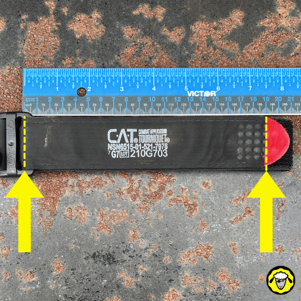 Garrot tourniquet CAT déplié à plat avec règle graduée en centimètres pour mesurer la longueur, flèches jaunes indiquant les points de pliage