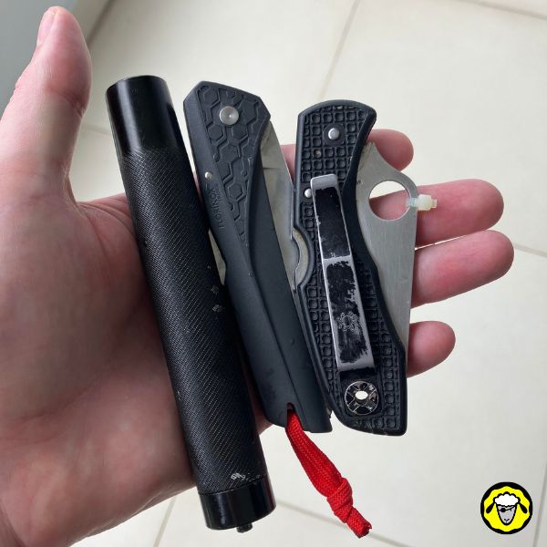 Main tenant une matraque télescopique compacte noire et deux couteaux pliants EDC sur un sol carrelé blanc.