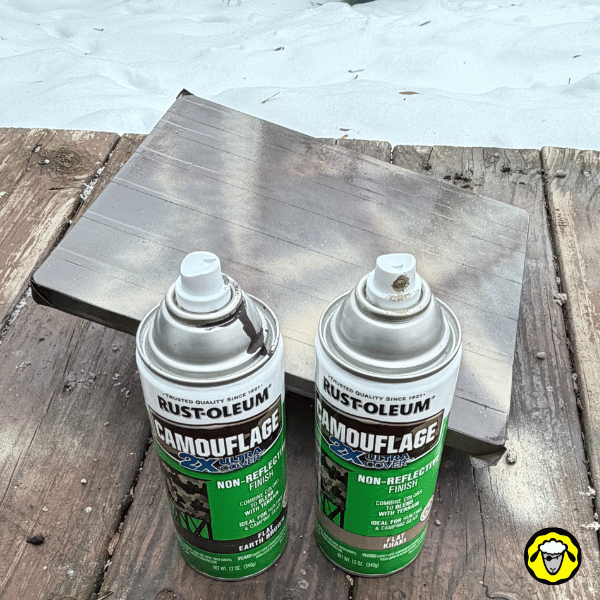 Deux bombes de peinture Rust-Oleum Camouflage posées devant une coque en silicone Starlink Mini présentant déjà un motif camouflage brun et beige, en cours de séchage sur une terrasse enneigée.