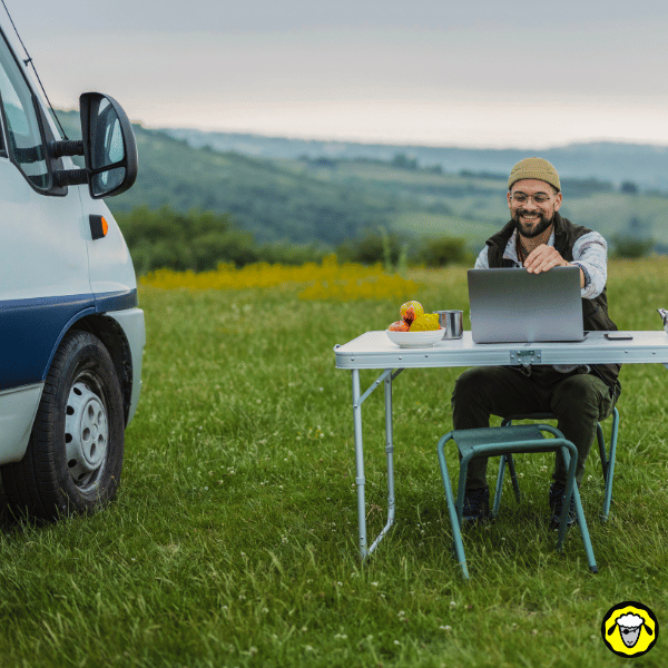 Homme assis à une table pliante près de son van, utilisant son ordinateur pour travailler en pleine nature grâce à une connexion haut débit mobile.