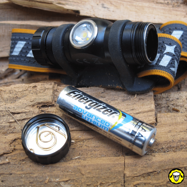 Lampe frontale Fenix HM23 avec compartiment ouvert et pile AA lithium à côté, posées sur du bois.