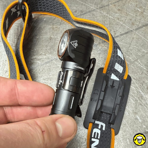 Lampe frontale Fenix HM23 posée avec son bandeau orange et le support de fixation, main montrant la lampe.