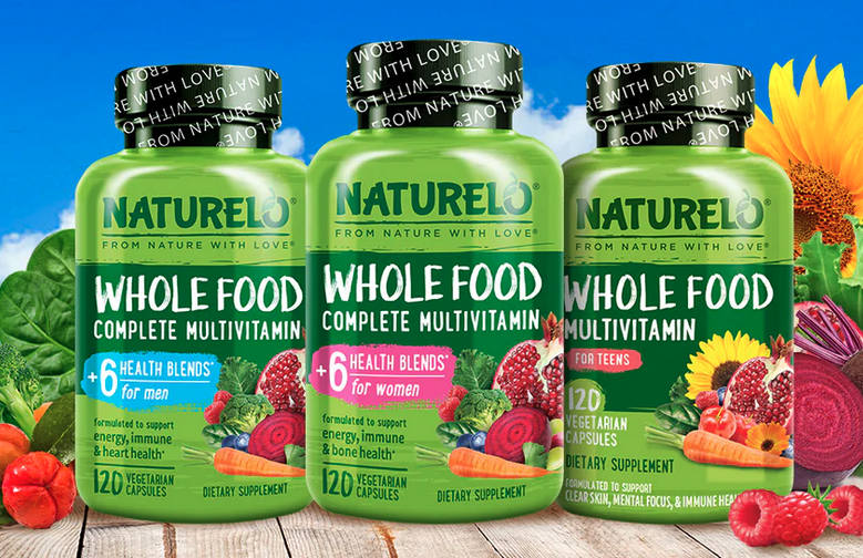 Trois flacons verts de compléments alimentaires Naturelo Whole Food Complete Multivitamin (hommes, femmes et adolescents) posés sur une table en bois avec fruits et légumes frais en arrière-plan sur fond bleu.