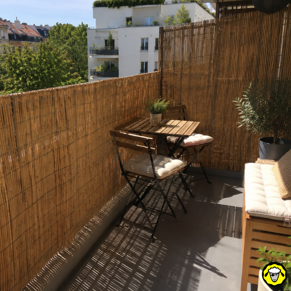 Balcon ombragé par des canisses, avec table, chaises et plantes en pot.